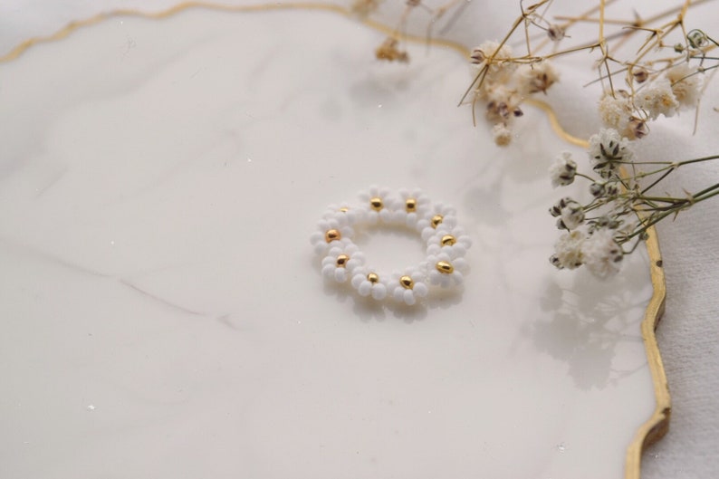 May include: A white beaded ring with gold accents, shaped like a daisy flower, on a white marble surface.