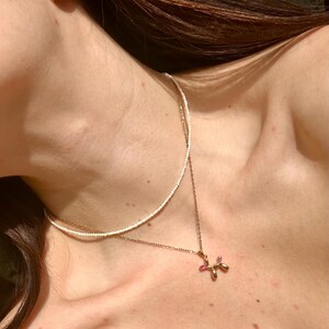 Puede incluir: Un collar de cadena dorada con un pequeño colgante de animal de globo rosa. El collar está superpuesto a un collar de cuentas blancas.