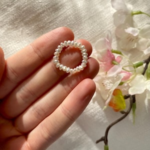 Puede incluir: Un anillo de perlas blancas hecho de pequeñas perlas redondas. El anillo está en una mano, y el fondo es una tela blanca con una flor rosa.