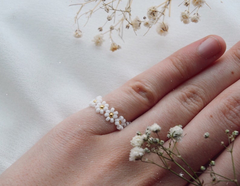 May include: A white daisy flower ring with gold accents. The ring is on a finger and the background is white.