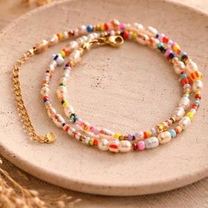 May include: A layered necklace with a gold chain and clasp, featuring a mix of white, colorful beads, and pearls. The necklace is displayed on a beige plate, with dried plants in the foreground.