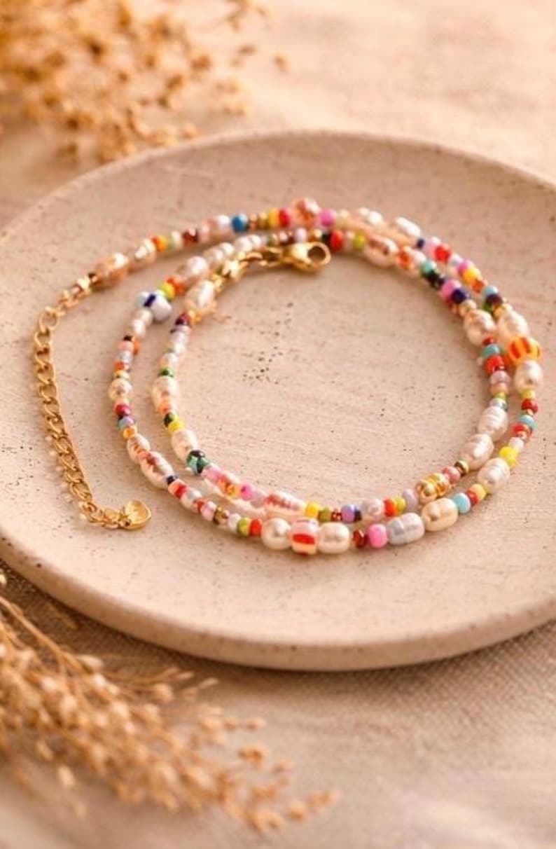 May include: A layered necklace with a gold chain and clasp, featuring a mix of white, colorful beads, and pearls. The necklace is displayed on a beige plate, with dried plants in the foreground.