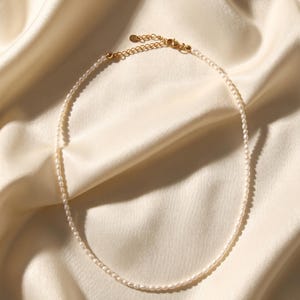 May include: Delicate pearl necklace with a gold-colored clasp and chain. The necklace is composed of small, round, white pearls. The jewelry is displayed on a cream-colored, silky fabric.