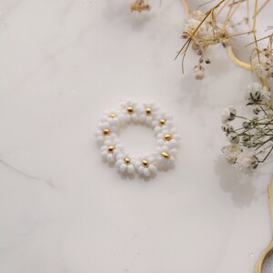 May include: A white daisy flower ring made of small beads with gold accents. The ring is on a white marble surface.