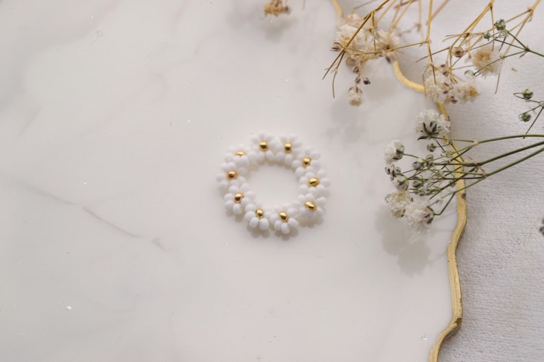 May include: A white daisy flower ring made of small beads with gold accents. The ring is on a white marble surface.