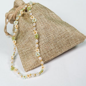 May include: A delicate necklace featuring freshwater pearls, small gold beads, and pastel-colored flower accents. The necklace is displayed next to a small, beige burlap drawstring bag, creating a rustic and elegant presentation. The necklace is a perfect accessory for any occasion.