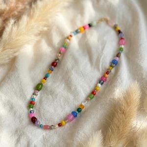 Könnte beinhalten: Eine farbenfrohe Perlenkette mit einer Vielzahl von Formen und Größen von Perlen. Die Perlen sind in den Farbtönen Rosa, Blau, Grün, Gelb und Rot gehalten. Die Kette liegt auf einer weißen Oberfläche mit getrockneten Pflanzen im Hintergrund.