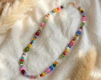 May include: A colorful beaded necklace with a variety of shapes and sizes of beads. The beads are in shades of pink, blue, green, yellow, and red. The necklace is on a white surface with dried plants in the background.