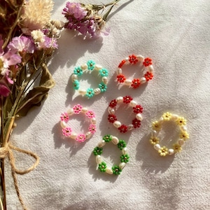 Könnte beinhalten: Sechs bunte Perlenringe mit perlmuttartigen Perlen und blütenförmigen Anhängern. Die Ringe sind in den Farben Türkis, Rot, Rosa, Gelb und Grün.