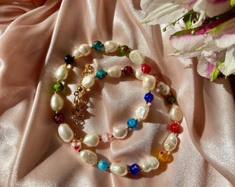 May include: A white pearl necklace with colorful glass beads. The necklace is on a pink satin fabric background.