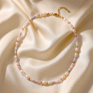 Könnte beinhalten: Eine zarte Halskette mit Süßwasserperlen, die mit kleinen goldenen Perlen und pastellfarbenen Perlen in Rosa und Blau durchsetzt sind. Die Halskette wird auf einer cremefarbenen Satin-Oberfläche präsentiert.
