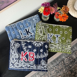 May include: Three rectangular quilted zipper pouches featuring paisley bandana patterns in light blue, olive green, and navy blue. Each pouch displays two large, contrasting fabric letters on the front: LK, KT, and KB, respectively.