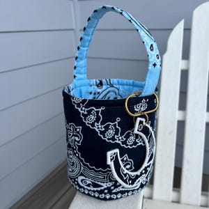 May include: A cylindrical fabric bucket with a handle, featuring a dark navy blue and white paisley bandana print. The interior is lined with light blue paisley fabric. A gold-toned metal ring holds a white embroidered letter C charm.
