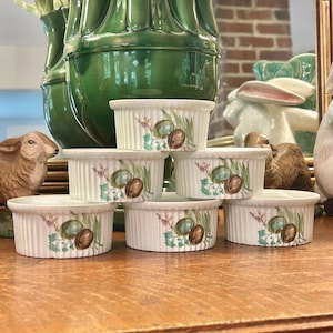 May include: A set of five white ceramic ramekins with vertical ribbed sides, each decorated with a botanical illustration featuring green leaves, small purple flowers, and two brown nuts. The small round dishes are arranged in a stacked formation.