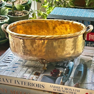 May include: An oval brass bowl with a hammered texture, featuring a twisted rope-style rim, two small side handles, and three decorative feet. The interior and exterior surfaces have a polished, reflective gold-toned metallic finish.