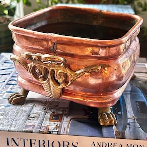 May include: A square copper planter with a metallic finish, featuring ornate gold-toned metal handles on the sides and four decorative claw-shaped feet. The vessel has a wide opening and a slightly rounded body with visible signs of patina.