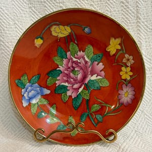 May include: A red ceramic plate with a gold rim and floral design. The plate features pink, blue, yellow, and green flowers on a red background.