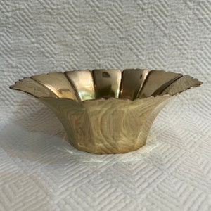 May include: A gold-colored metal bowl with a scalloped edge and a fluted design. The bowl is sitting on a white textured surface.