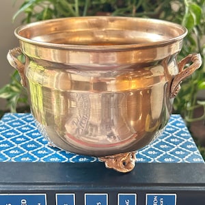 May include: A shiny, gold-toned metal planter with two ornate handles and decorative feet. The planter sits atop a stack of books with titles including "Reader's Digest Condensed Books" and "Lady Washington". Green foliage is visible in the background.