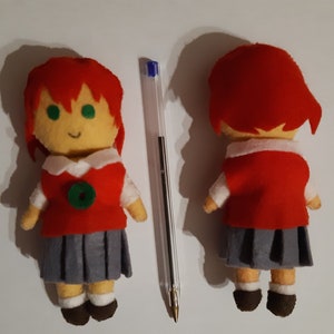 Peut inclure: Un jouet en peluche fait main représentant une fille aux cheveux roux, portant un pull rouge avec un cercle vert sur le devant, une jupe grise et des chaussettes blanches. Le jouet est debout sur une surface blanche.