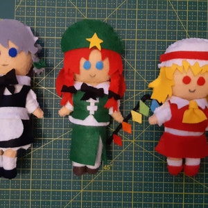 May include: Three handmade felt dolls with detailed features. The dolls have different colored hair, outfits, and accessories. The doll on the left has gray hair and a black and white outfit. The middle doll has red hair and a green outfit. The doll on the right has yellow hair and a red outfit.