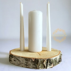 Può includere: Un portacandele rustico con una base in legno naturale e tre candele bianche. La composizione comprende due candele affusolate alte e sottili e una candela a colonna cilindrica più corta. La base in legno è di colore marrone chiaro.