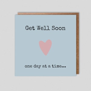 Puede incluir: Una tarjeta azul claro con el texto "Get Well Soon" en fuente negra. Un corazón rosa está en el centro de la tarjeta. El texto "one day at a time..." está debajo del corazón.