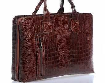 May include: A brown leather briefcase with a crocodile-embossed pattern. The briefcase has a zipped pocket on the front and two top handles.