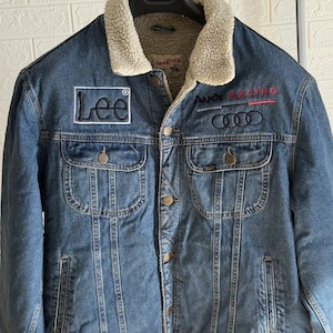 May include: A blue denim jacket with a sherpa lining. The jacket has a Lee logo patch on the left chest and an Audi Racing logo on the right chest. The jacket has a button closure and two front pockets.