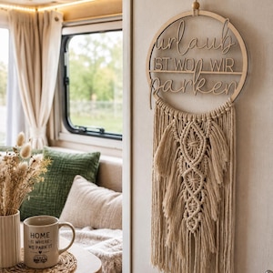 Könnte beinhalten: Ein runder Wandbehang aus Holz mit dem deutschen Schriftzug "urlaub IST WO WIR parken" in einem Ring, darunter beigefarbene Makramee-Fransen. Daneben steht eine Keramiktasse mit dem Text "HOME IS WHERE WE PARK IT" und einem Wohnmobil-Symbol.