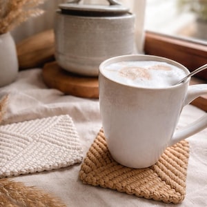 Könnte beinhalten: Eine weiße Keramiktasse mit einem schaumigen Getränk steht auf einem beigefarbenen gewebten Untersetzer. Ein zweiter, ähnlicher Untersetzer ist zu sehen, zusammen mit einem kleinen Keramiktopf und getrockneten Pflanzen. Die Szene spielt auf einer neutralen Leinenoberfläche.