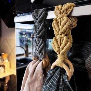 Könnte beinhalten: Zwei Makramee-Küchenhandtuchhalter in Grau und Beige, die an einem Ofengriff hängen. Jeder Halter hat ein geflochtenes Seildesign und einen Holzring. Die Handtücher sind hellrosa und dunkelgrau kariert.