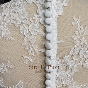 A white lace fabric with a row of white buttons down the center back. The lace has a floral pattern. Beautiful wedding dress topper, Alecon Lace bridal topper, handcrafted in Florida. Highe neck, long sleeves and cover buttons. A white lace wedding dress bodice with a high neck and long sleeves. The lace is delicate and features floral patterns.