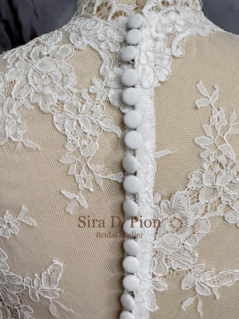 A white lace fabric with a row of white buttons down the center back. The lace has a floral pattern. Beautiful wedding dress topper, Alecon Lace bridal topper, handcrafted in Florida. Highe neck, long sleeves and cover buttons. A white lace wedding dress bodice with a high neck and long sleeves. The lace is delicate and features floral patterns.