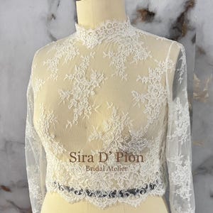 May include: A sheer, ivory lace top with long sleeves and a high neckline. The delicate floral lace pattern covers the entire top, with a scalloped edge at the waist. The top is displayed on a mannequin.