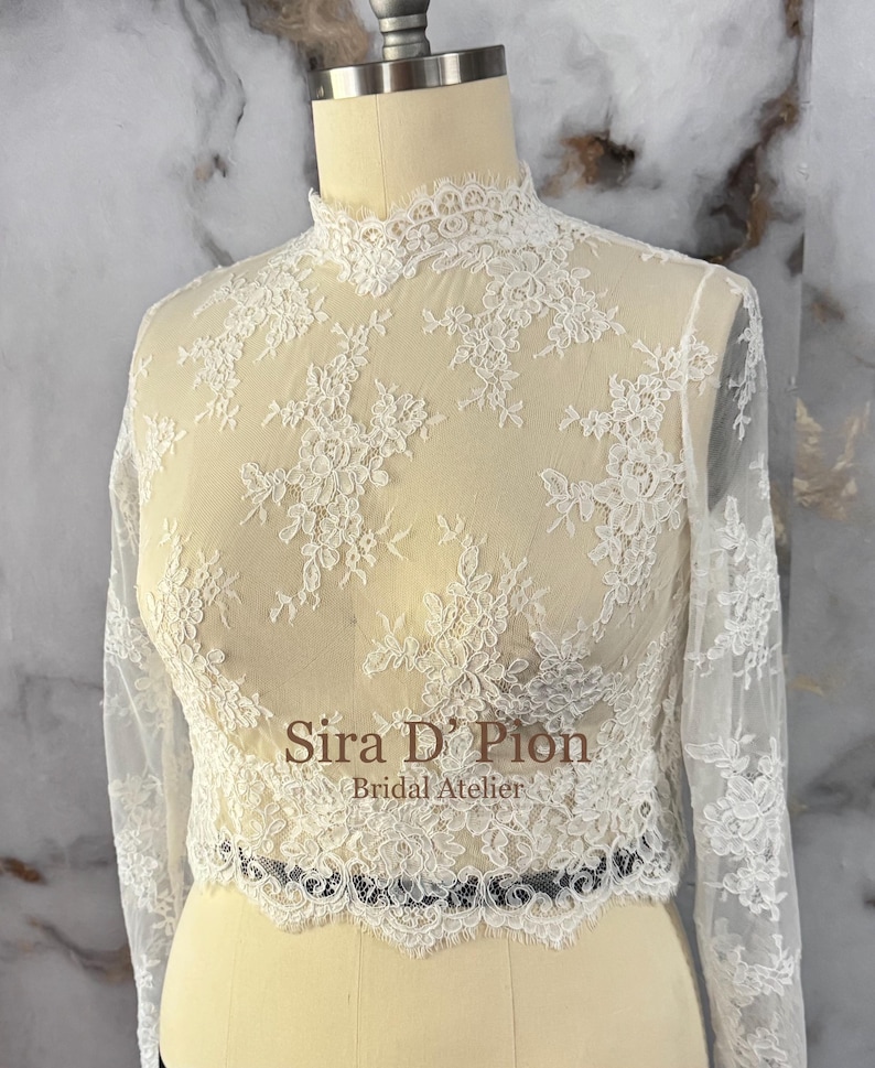 May include: A sheer, ivory lace top with long sleeves and a high neckline. The delicate floral lace pattern covers the entire top, with a scalloped edge at the waist. The top is displayed on a mannequin.