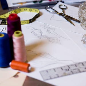 May include: A close-up of a sewing project with spools of thread in various colors, including blue, pink, red, and orange. A measuring tape, a pair of scissors, and a ruler are also visible. The project appears to be a garment design.