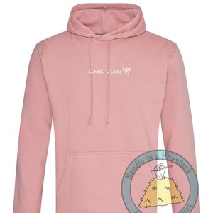 May include: A pink hoodie with a white graphic that says "Good Vibes" and a hand drawn hang loose symbol. The hoodie has a kangaroo pocket and a drawstring hood.