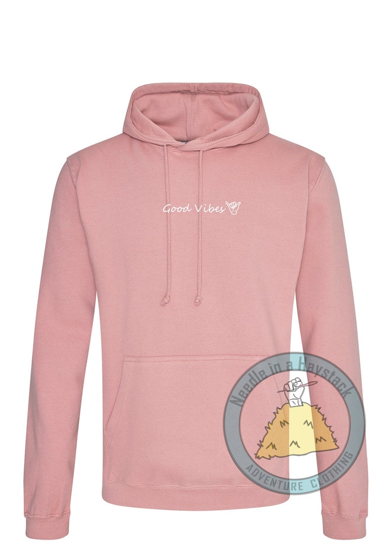 May include: A pink hoodie with a white graphic that says "Good Vibes" and a hand drawn hang loose symbol. The hoodie has a kangaroo pocket and a drawstring hood.