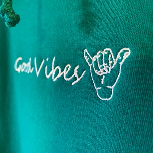 May include: A teal green hoodie with white embroidered text that says "Good Vibes" and a white embroidered hand gesture.