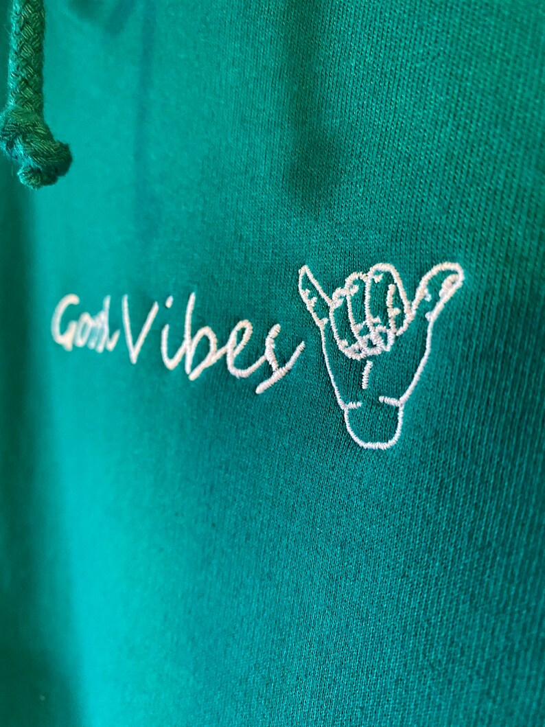 May include: A teal green hoodie with white embroidered text that says "Good Vibes" and a white embroidered hand gesture.