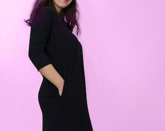May include: A black, knee-length dress with three-quarter sleeves and pockets. The dress has a relaxed fit and a slightly curved hemline. The background is a solid pink colour.