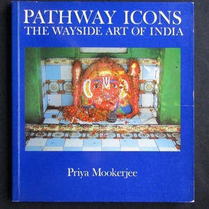 Könnte beinhalten: Ein quadratisches Buch mit dem Titel Pathway Icons: The Wayside Art of India von Priya Mookerjee. Der blaue Einband zeigt ein zentrales Foto einer leuchtend orangefarbenen Gottheitsstatue mit großen Augen in einem gefliesten Schrein mit Blumenschmuck.