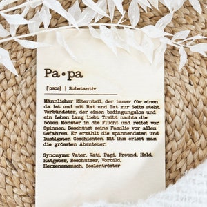 May include: A wooden plaque with the German word "Papa" and its definition. The plaque has text in black font and is decorated with white floral accents. The background is a woven texture.