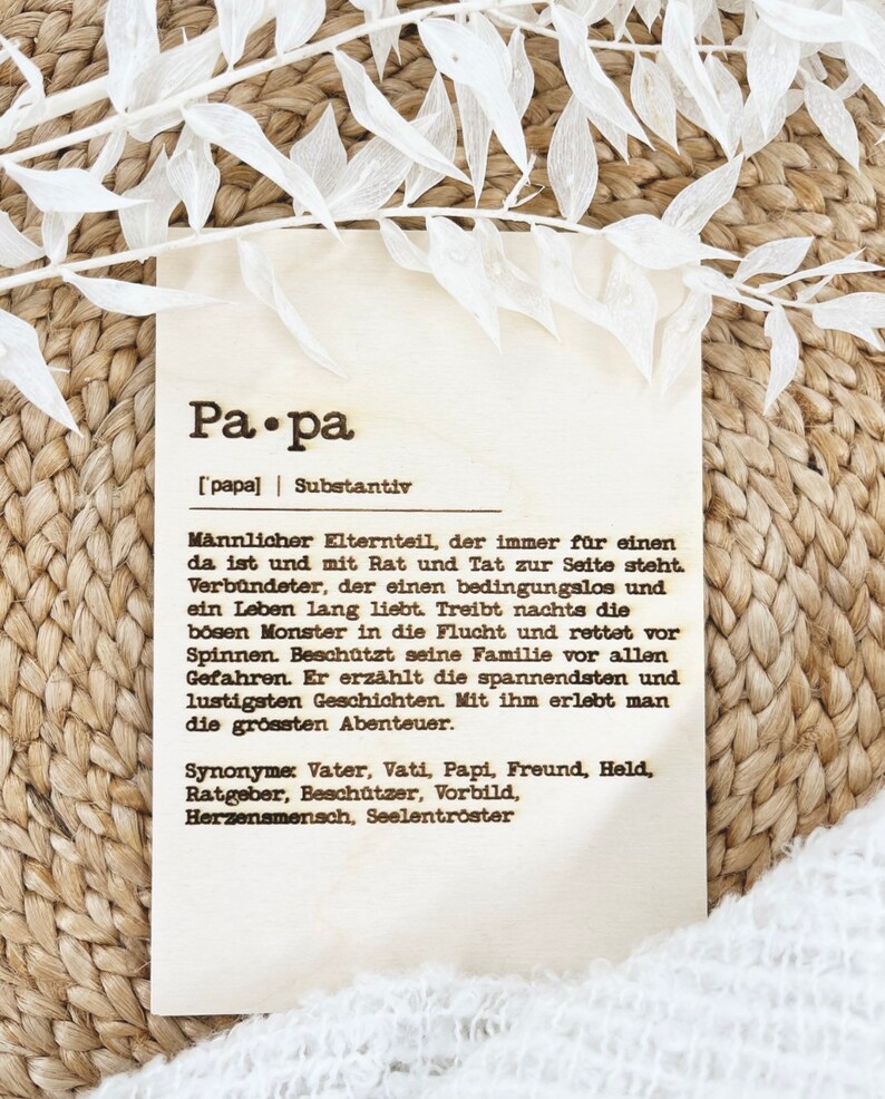 May include: A wooden plaque with the German word "Papa" and its definition. The plaque has text in black font and is decorated with white floral accents. The background is a woven texture.