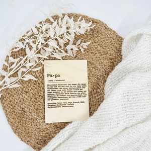 May include: A wooden plaque with the word "Pa.pa" and a definition in German, resting on a woven placemat. A white sprig of leaves and a white, textured cloth are also in the image. The plaque is a decorative item.