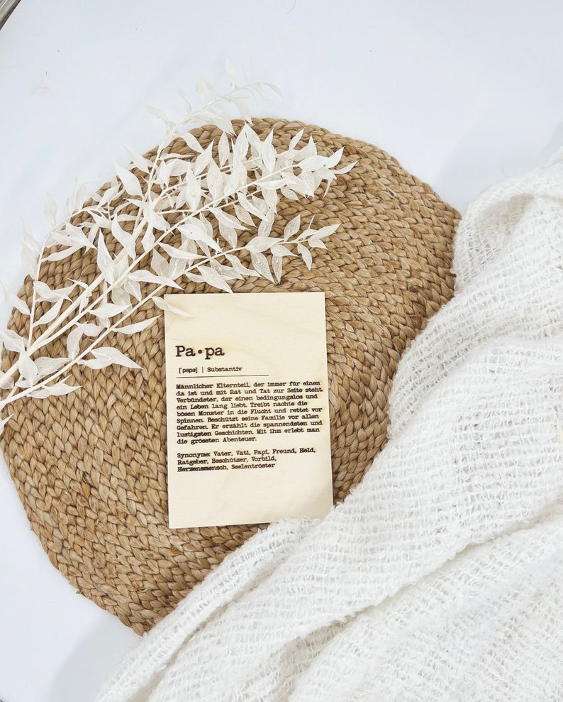 May include: A wooden plaque with the word "Pa.pa" and a definition in German, resting on a woven placemat. A white sprig of leaves and a white, textured cloth are also in the image. The plaque is a decorative item.