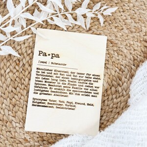 May include: A rectangular wooden plaque with the German word "Papa" and its definition printed in black. The plaque is on a woven surface with white decorative leaves.