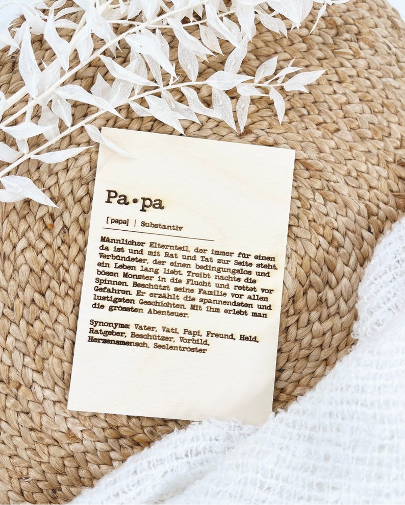 May include: A rectangular wooden plaque with the German word "Papa" and its definition printed in black. The plaque is on a woven surface with white decorative leaves.