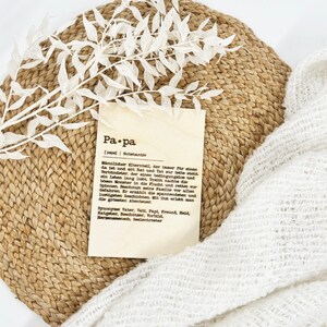 May include: A decorative arrangement featuring a woven placemat, a sprig of white leaves, a cream-colored fabric, and a card with the word "Pa.pa" and German text. The card is a definition of the word "father".